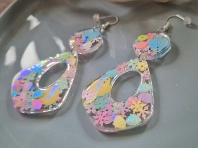 In den transparenten großzügen Boho-Anhängern wurden glitzernde Blumen- und Muschel-Pailletten in sommerlichen Pastelltönen gegossen, die je nach Lichteinfall strahlend türkis-changierend leuchten, Akzente setzen und diese Statementohrringe zu  individuellen Begleitern machen.  Echte handgefertigte Kunstohrringe