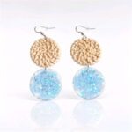 Unter dem hellbeigen Rattankreis baumeln zart gegossene kreisrunde transparente Anhänger mit kleinen glitzernden hellblauen Sternchen, die je nach Lichteinfall changieren.