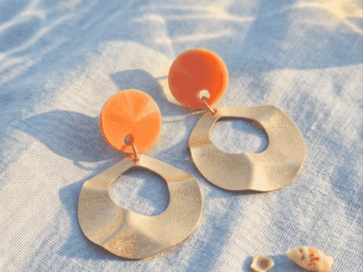 Statement-Ohrringe „Summer Wave“ mit orangem Highlight & GoldSpüre die Wärme der Sonne und das Rauschen des Meeres. Diese ausdrucksstarken Hängeohrringe kombinieren ein kräftiges Orange mit matt-goldenen Elementen, deren wellige Struktur an sanfte Meeresbewegungen erinnert. Perfekt inszeniert im warmen Sonnenlicht, bringen sie Frische und Urlaubsflair in jeden Look. Kombiniere sie mit einem luftigen Leinenkleid für den perfekten Beach-to-Dinner-Style.