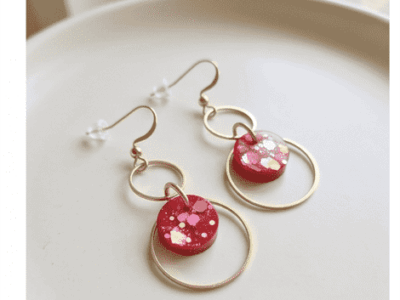 Kunst für deine Ohren! Diese dreistufigen Statement-Ohrhänger vereinen minimalistische Geometrie mit einem aufregenden Farbakzent. Die handgefertigten rot-glitzernden Designkreise bestichen durch ihre einzigartigen, eingearbeiteten Farb- und Metallic-Sprenkel, die an modernes Terrazzo-Muster erinnern. Eingebettet in zwei ineinandergreifende, goldfarbene Ringe entsteht ein dynamischer Look, der bei jeder Bewegung sanft mitschwingt. Ein echtes Unikat für Individualistinnen.