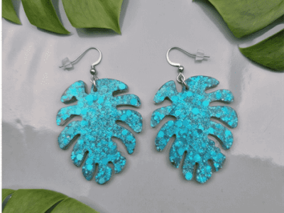 Paradiesische Vibes f&uuml;r dein Outfit! Die ausdrucksstarken H&auml;ngeohrringe fangen das Licht bei jeder Bewegung ein. Die filigranen Monstera-Bl&auml;tter sind gef&uuml;llt mit unz&auml;hligen t&uuml;rkisfarbenen Glitzerpartikeln und feinen Flakes, die dem Schmuckst&uuml;ck eine wundersch&ouml;ne optische Tiefe verleihen.