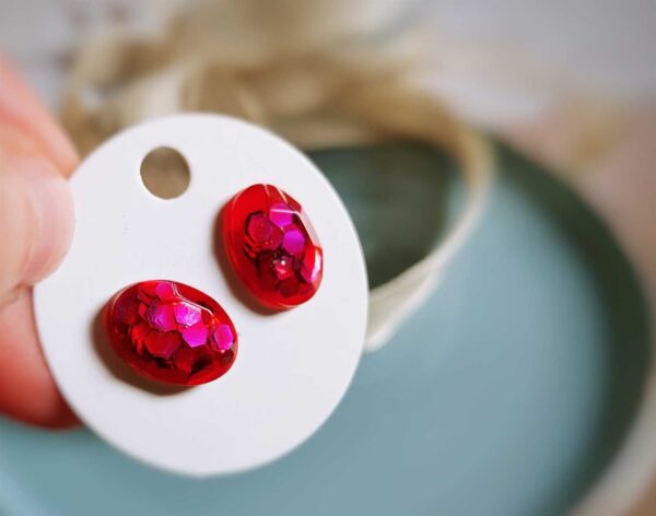 Diese handgefertigten oval-kristallförmigen poppigen Ohrstecker setzen auf eine mondäne Weise Akzente und überzeugen durch ein transparentes Rot mit knalligen pinken Glitzer-Pailletten.
